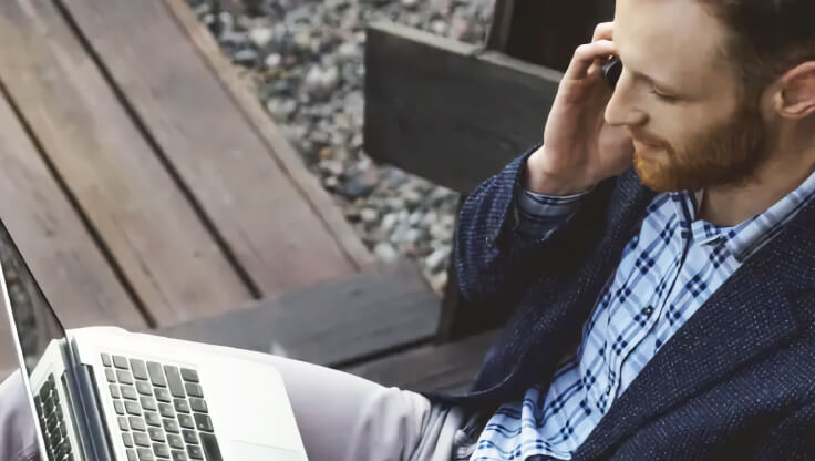 A man is working using his mobile phone and laptop on a bench