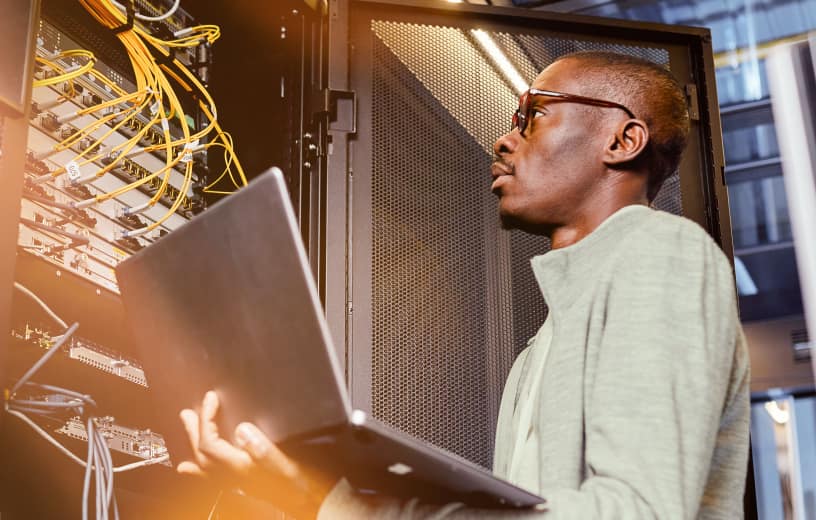 An IT guy holding a laptop while looking at a server with multiple cables